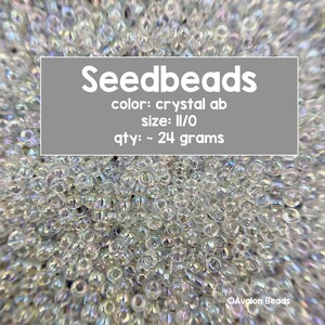 May include: A close-up of a pile of clear seed beads with an iridescent finish. The beads are labeled "Seedbeads", "color: crystal ab", "size: II/O", and "qty: ~ 24 grams".