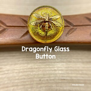 Puede incluir: Un botón de vidrio amarillo con un diseño de libélula dorada. El botón tiene una superficie texturizada y es perfecto para agregar un toque de fantasía a cualquier proyecto.