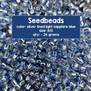 May include: A close-up shot of a collection of seed beads. The beads are silver-lined light sapphire blue, size 8/0. The image includes the text "Seedbeads" and "qty: ~ 24 grams".