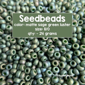 Pode incluir: Um close-up de um monte de contas de sementes de cor verde-sálvia fosco com brilho, tamanho II/O. As contas pesam aproximadamente 24 gramas. O texto "Seedbeads" está escrito em branco sobre um retângulo preto, com as palavras "color: matte sage green luster", "size: II/O" e "qty: ~ 24 grams" escritas abaixo. O texto "Avalon Beads" está escrito em branco sobre um retângulo preto no canto inferior direito da imagem.