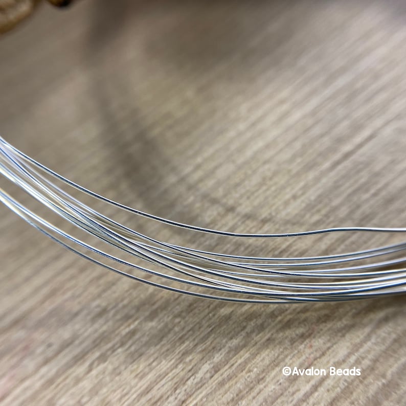 May include: Close-up of several strands of silver-colored wire, likely for jewelry making or crafts. The wire is thin and flexible, with a slight curve. The background is a light brown wooden surface. The text "Avalon Beads" is in the bottom right corner.