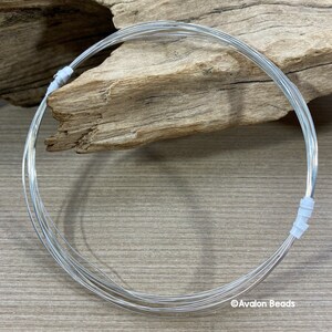 May include: A coil of bright silver-colored wire, likely for jewelry making or crafts, is neatly arranged in a circular shape. The wire is held together with small white plastic pieces. The background features a piece of driftwood on a wooden surface.