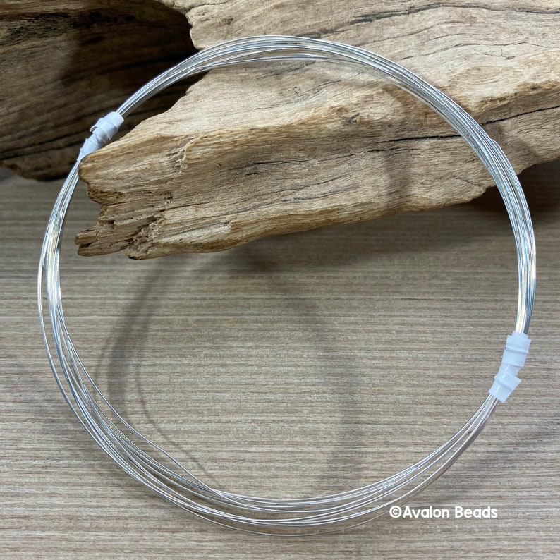 May include: A coil of bright silver-colored wire, likely for jewelry making or crafts, is neatly arranged in a circular shape. The wire is held together with small white plastic pieces. The background features a piece of driftwood on a wooden surface.