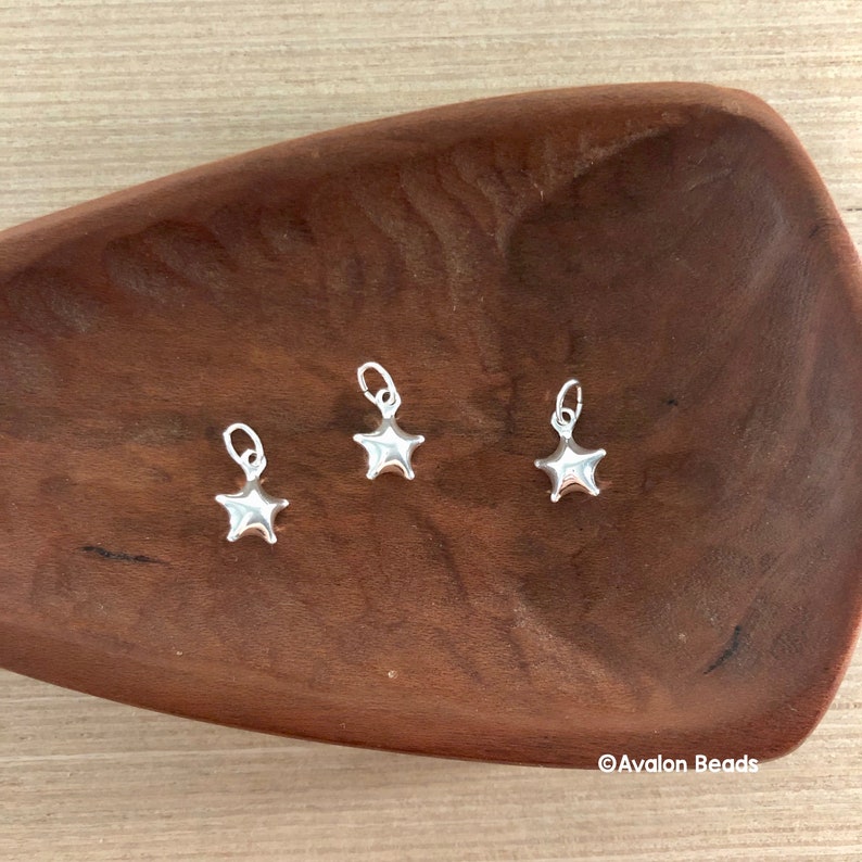 May include: Three silver star-shaped charms on a wooden surface. The charms have a smooth, shiny finish and are perfect for adding a touch of sparkle to jewelry projects.