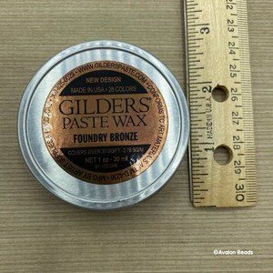 May include: A silver tin of Gilders Paste Wax in Foundry Bronze. The tin is labeled with the brand name, product name, color, and instructions for use. It covers 30 square feet and contains 1 ounce (30 ml) of wax.