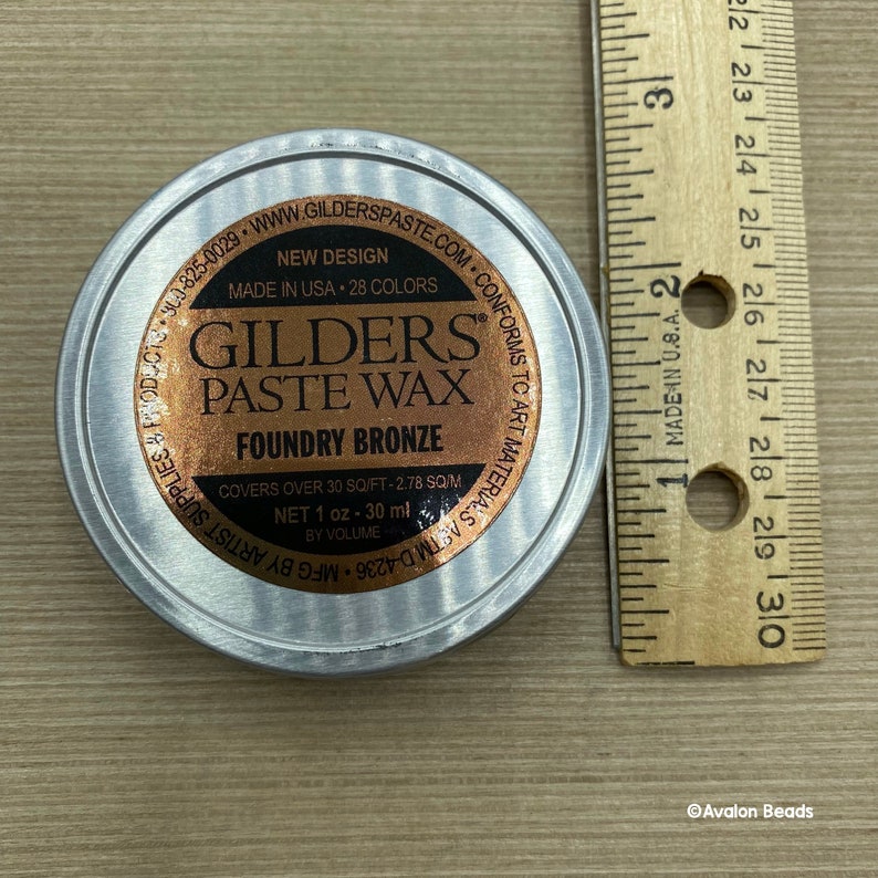 May include: A silver tin of Gilders Paste Wax in Foundry Bronze. The tin is labeled with the brand name, product name, color, and instructions for use. It covers 30 square feet and contains 1 ounce (30 ml) of wax.