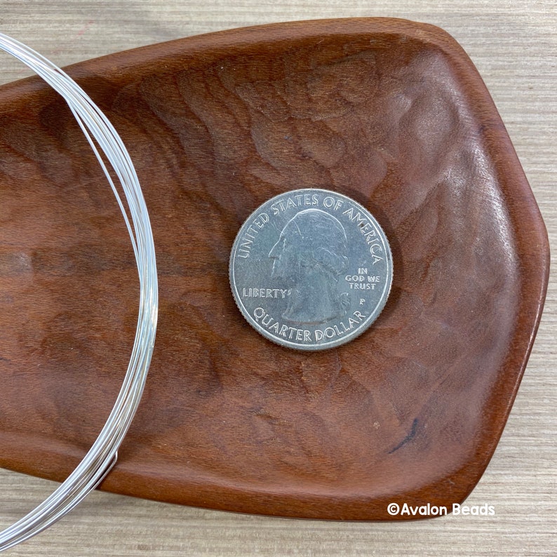 May include: A quarter coin next to a coil of silver-colored wire, displayed on a dark wooden tray. The coin shows the profile of George Washington and the words "Quarter Dollar". The wire is tightly wound, suggesting it is for crafting or jewelry making.