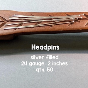 May include: A collection of silver-filled headpins resting on a carved wooden surface. The text reads: Headpins, silver filled, 24 gauge, 2 inches, qty. 50. These thin, straight metal pins feature a small flat head at one end for jewelry making.