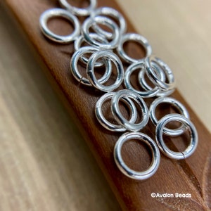 May include: A close-up of a pile of silver jump rings on a wooden surface. The rings are all the same size and are arranged in a random pattern. The rings are shiny and have a smooth finish.
