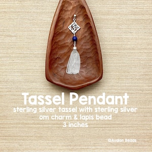 May include: A 3-inch sterling silver tassel pendant featuring a silver Om charm and a single round blue lapis bead. The pendant is displayed inside a carved wooden bowl. The text on the image reads Tassel Pendant, sterling silver tassel with sterling silver om charm & lapis bead, 3 inches, and Avalon Beads.