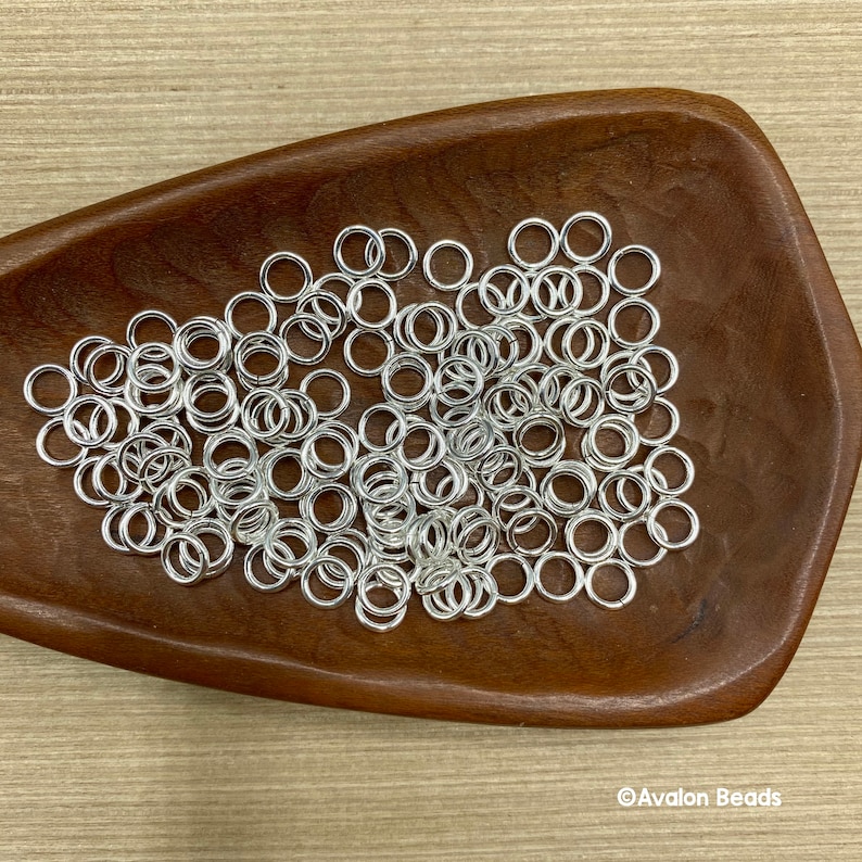 May include: A close-up image of a wooden bowl filled with silver jump rings. The jump rings are small and round, and they are all piled together in a heap.
