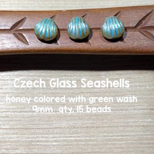 May include: Three Czech glass seashell beads. The honey-colored beads have a green wash and blue stripes. The text on the image reads "Czech Glass Seashells honey colored with green wash 9mm qty. 15 beads."