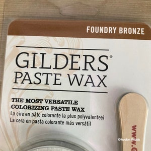 May include: A container of Foundry Bronze GILDERS PASTE WAX, a versatile coloring paste wax. The label reads "The Most Versatile Coloring Paste Wax" and includes French and Spanish translations. The container is accompanied by a wooden applicator.