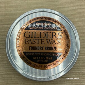 May include: A round metal container with a label that reads "GILDERS PASTE WAX FOUNDRY BRONZE" in gold lettering. The label also states "NET 1 oz - 30 ml BY VOLUME" and "COVERS OVER 30 SQ/FT - 2.78 SQ/FT".
