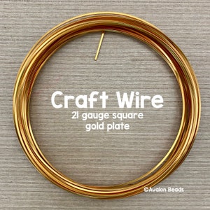 May include: A coil of gold-plated 21 gauge square craft wire. The wire is wound in a circle and is on a wooden surface.