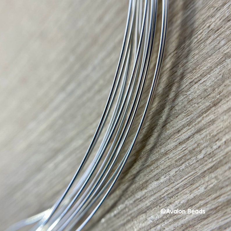 May include: Close-up of several strands of shiny, silver-colored wire, likely for jewelry making or crafts. The wires are neatly arranged and slightly curved, with a wooden surface in the background. The Avalon Beads logo is visible in the bottom right corner.