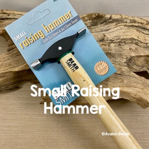 Può includere: Un piccolo martello da battitura con manico in legno, testa in metallo nero e dettagli verdi. Il martello ha due facce, una con una superficie di 2x13mm e l'altra con una superficie di 6x13mm. La confezione riporta "Piccolo martello da battitura" e "Bead Smith" con un adesivo "Qualità Pro".