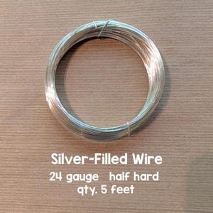 Può includere: Una bobina di filo riempito d'argento, calibro 24, semirigido. Il testo sull'immagine recita "Silver-Filled Wire 24 gauge half hard qty. 5 feet". Il filo è arrotolato, probabilmente per la creazione di gioielli o progetti artigianali. Circa 1,52 metri.