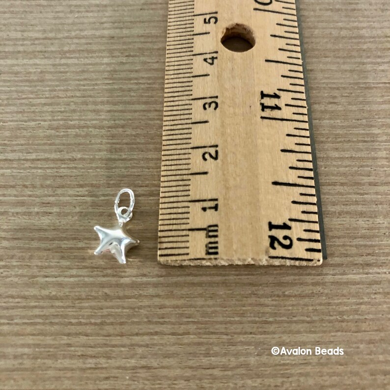 May include: A silver star charm measuring approximately 1/2 inch in diameter. The charm is shown next to a ruler for size reference.