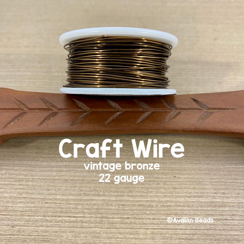 May include: A spool of vintage bronze craft wire in 22 gauge. The wire is wound around a white plastic spool. The spool is sitting on a wooden surface.