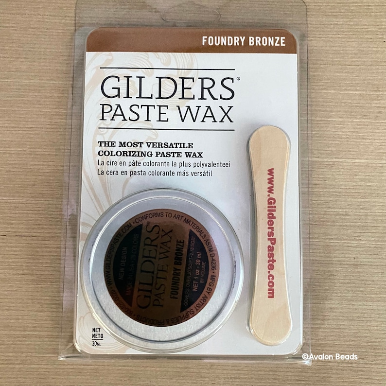 May include: A round tin of Gilders Paste Wax in Foundry Bronze. The tin is labeled with the product name, color, and website address. The tin is packaged with a wooden applicator stick.