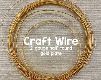 Puede incluir: Un rollo de alambre artesanal de calibre 21, medio redondo, chapado en oro. El alambre está enrollado en círculo sobre una superficie de madera. El texto "Craft Wire 21 gauge half round gold plate" está impreso en la imagen.