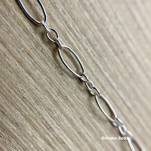 May include: Close-up of a silver chain with oval links. The chain is made of shiny, polished metal and is displayed against a light brown wooden surface. The chain links are connected by small, round rings.