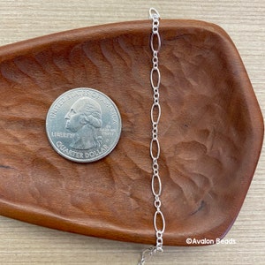 May include: A silver chain with oval links is displayed next to a quarter on a wooden surface. The chain is delicate and shiny, with a repeating pattern of elongated oval shapes. The quarter provides a size reference.