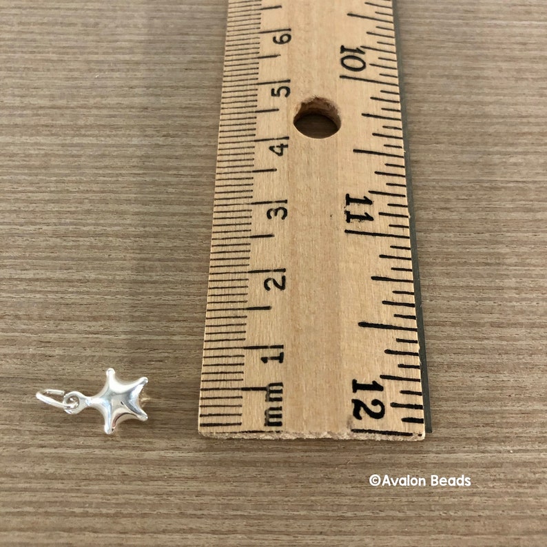 May include: A silver star charm measuring approximately 1/2 inch in diameter. The charm is shown next to a ruler for size reference.