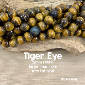 Könnte beinhalten: Eine Strähne aus 12 mm runden Tigeraugenperlen mit einem großen 4 mm Loch. Die Perlen sind in einem Mix aus goldbraunen und dunkelbraunen Farben. Die Perlen liegen auf einer Holzoberfläche. Tiger Eye 12mm rund großes 4mm Loch Menge: 1 Strang ©Avalon Beads