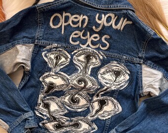 Puede incluir: Chaqueta vaquera azul oscuro con mangas plateadas y diseño pintado a mano. La parte trasera de la chaqueta presenta las palabras "open your eyes" sobre una serie de ilustraciones de ojos estilizados en blanco y negro.