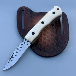 May include: A hunting knife with a cream-colored handle and a patterned blade rests on a brown leather sheath. The handle has black rivets. The sheath has a decorative embossed design.