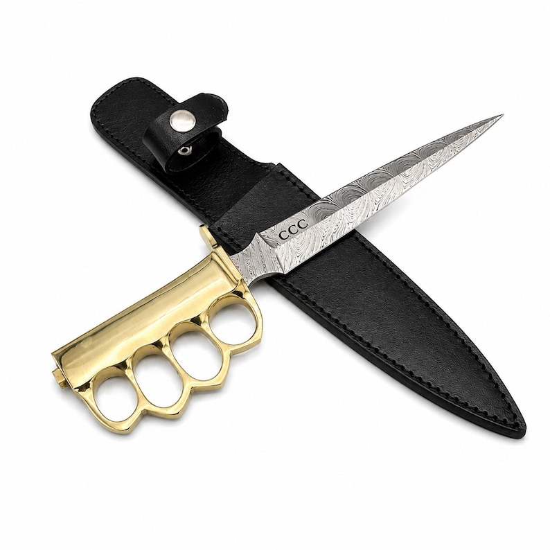 May include: A fixed-blade knife with a polished gold-tone brass knuckle handle and a pointed Damascus steel blade featuring a swirling pattern. The blade is engraved with the letters CCC. It is accompanied by a black leather sheath with a snap closure.
