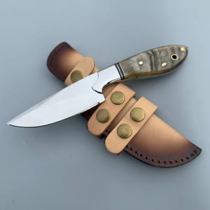 Puede incluir: Un cuchillo artesanal con una hoja de acero pulido y un mango con un patrón marrón claro y gris. El cuchillo está asegurado en una funda de cuero color canela con cierres de botón color latón. El diseño sugiere un cuchillo de caza o utilitario.