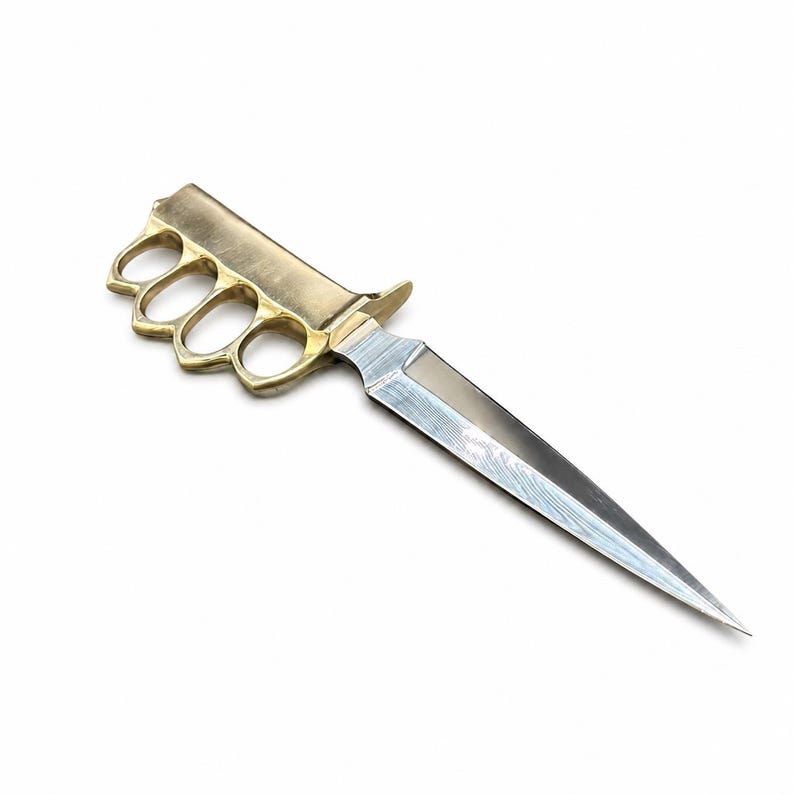 May include: A trench knife featuring a brass knuckle handle with four finger holes and a double-edged steel blade. The blade displays a distinct wavy pattern along its surface, while the handle has a smooth, metallic gold-toned finish.