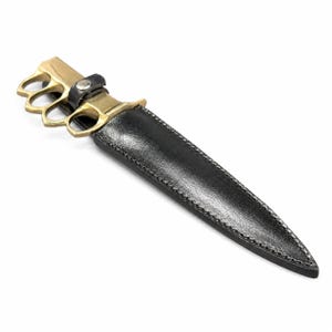 May include: A fixed-blade knife featuring a brass knuckle-style handle and a metallic bolster. The blade is encased in a black sheath with visible stitching along the edges and a small black strap secured with a silver-toned metal snap button.