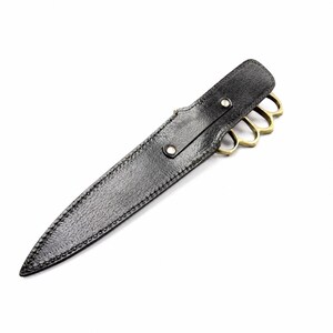 May include: A black textured sheath with visible stitching and two silver-toned metal rivets, housing a knife with a brass-colored knuckle duster handle. The handle features four finger holes and a metallic finish, partially visible from the sheath.