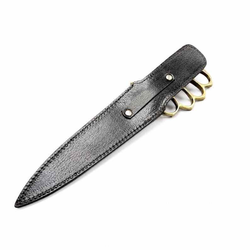 May include: A black textured sheath with visible stitching and two silver-toned metal rivets, housing a knife with a brass-colored knuckle duster handle. The handle features four finger holes and a metallic finish, partially visible from the sheath.