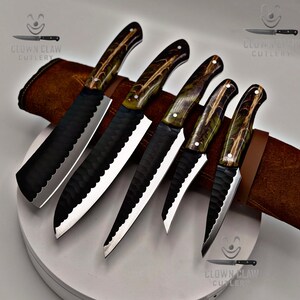 May include: A set of five kitchen knives with black blades and patterned wooden handles. The blades have a textured design and are displayed with a brown leather sheath. The knives are arranged on a white surface.