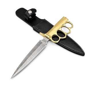 May include: A dagger with a patterned Damascus steel blade featuring the letters CCC, a gold-colored brass knuckle handle, and a black leather sheath with a snap button closure. The blade is pointed and double-edged with a distinct wavy texture.