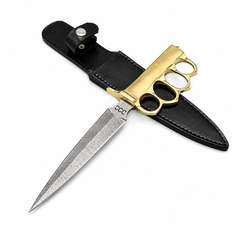 May include: A dagger with a patterned Damascus steel blade featuring the letters CCC, a gold-colored brass knuckle handle, and a black leather sheath with a snap button closure. The blade is pointed and double-edged with a distinct wavy texture.