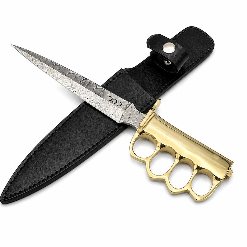 May include: A fixed-blade knife with a Damascus steel blade featuring the letters CCC, a polished gold-tone brass knuckle handle, and a black leather sheath with a snap button closure. The blade has a distinct wavy pattern throughout its surface.