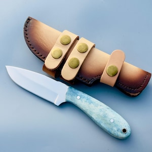 May include: A handcrafted knife with a light blue and white handle and a silver blade. The knife is paired with a tan leather sheath with brass-coloured button closures. The sheath has a dark brown border.