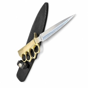 May include: A trench knife featuring a double-edged Damascus steel blade and a brass knuckle handle. The knife is positioned over a black leather sheath with a snap closure strap. The blade has a visible pattern and a metallic finish.