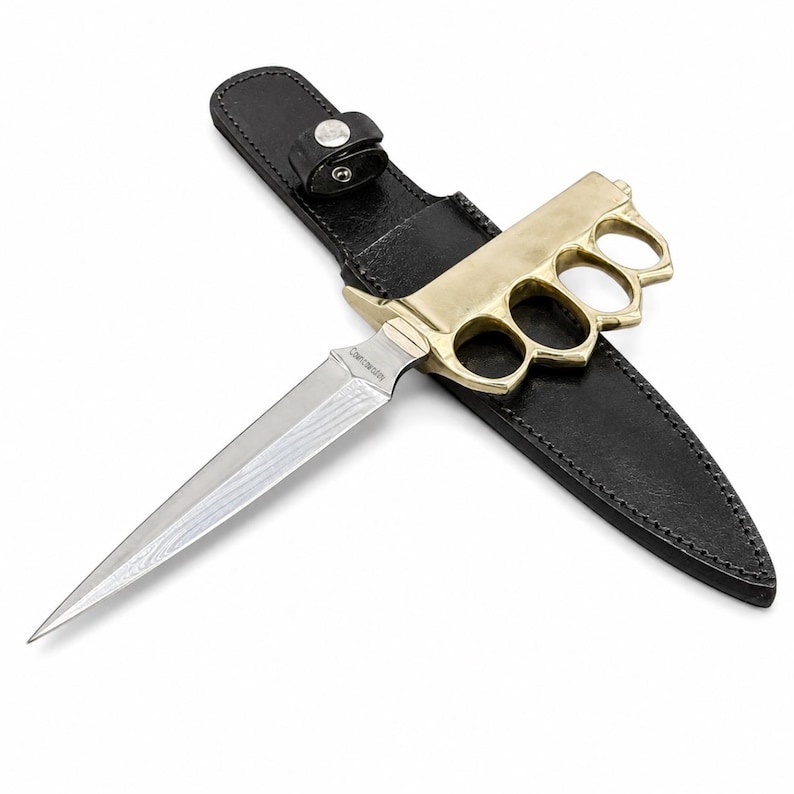 May include: A double-edged dagger with a Damascus steel blade and a brass knuckle-duster handle. The knife rests on a black leather sheath featuring a snap-button strap. The blade displays a visible wavy pattern, and the handle has four finger holes.