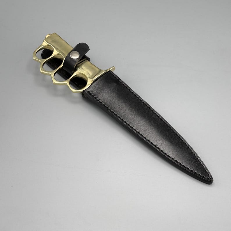 May include: A fixed-blade knife featuring a polished brass knuckle-duster handle and a black leather sheath. The sheath is secured with a black leather strap and a silver-toned metal snap button, covering the blade while exposing the brass handle.