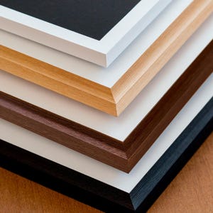May include: Close-up of a stack of picture frames in various colors. The frames include white, light brown, dark brown, and black. Each frame has a beveled edge and a white interior. The frames are stacked diagonally, showcasing their different colors and textures.