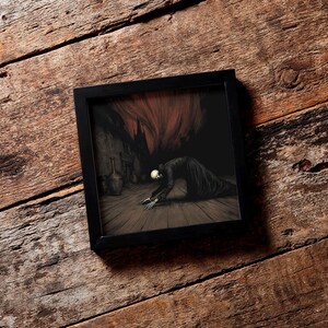 May include: A framed artwork depicting a skeletal figure in a dark robe kneeling on a wooden floor. The background features a shadowy interior with a fiery, red-orange element above. The frame is black.