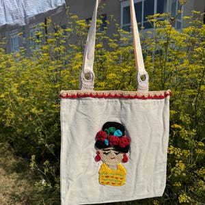 Puede incluir: Un bolso tote de color crema con un diseño inspirado en Frida Kahlo. El bolso presenta una imagen de ganchillo de una mujer con un sombrero negro adornado con flores rojas, detalles azules y ropa amarilla. Tiene asas color crema y un ribete de ganchillo rojo.