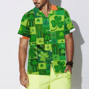 May include: Green button-up shirt with a patchwork design featuring shamrocks in various shades of green. The shirt has short sleeves with white cuffs and an orange collar. The shirt is worn with yellow shorts.